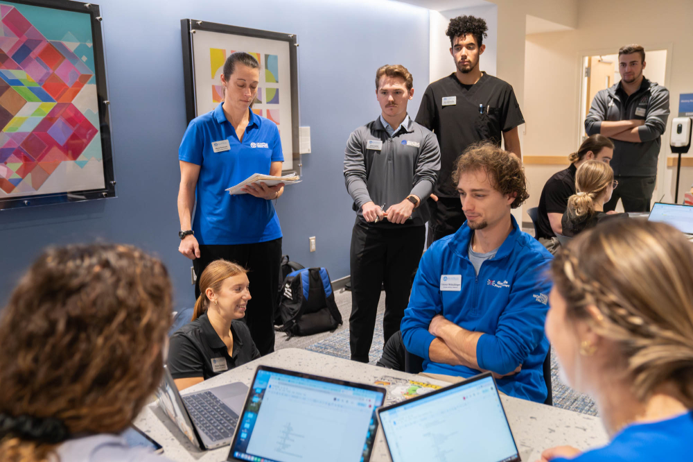 seven students, some seated in front of laptops, some standing in hallway