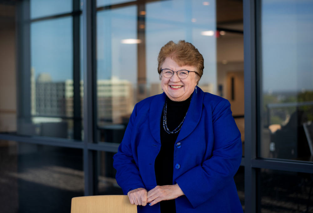 Linda Lewandowski in a blue jacket and black shirt stands with hands resting on a tall chair