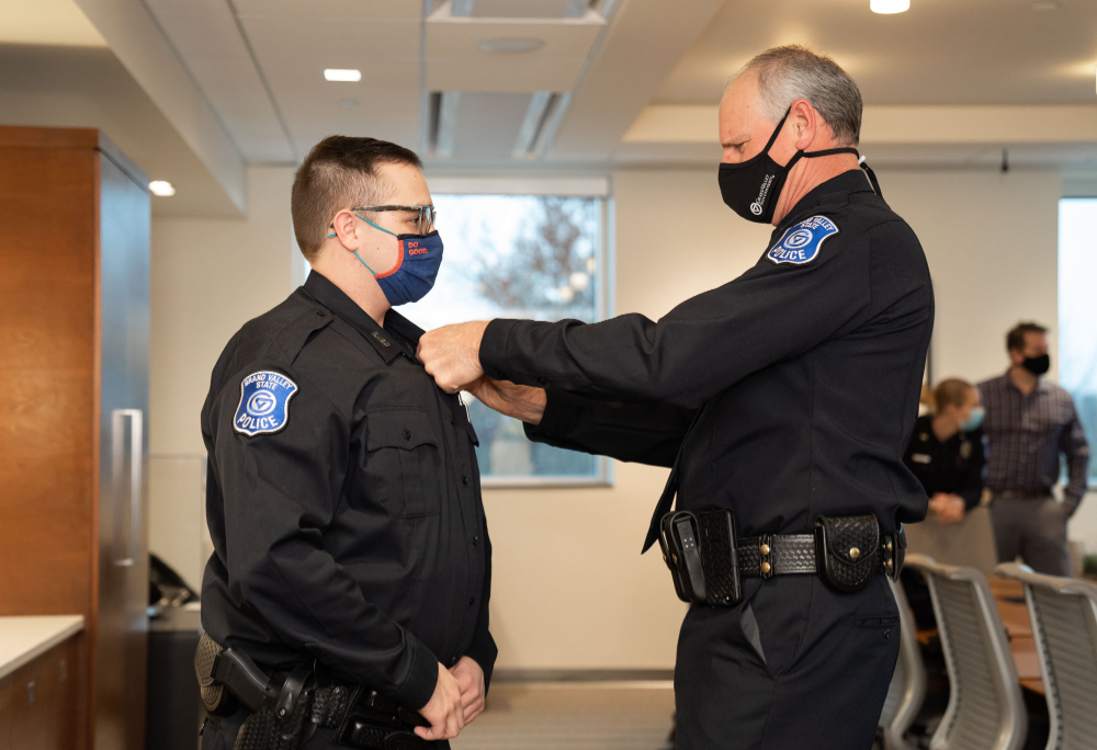 Brendan Estlick receives a pin from Brandon DeHaan, both are inside a room and wearing masks and GVPD uniforms