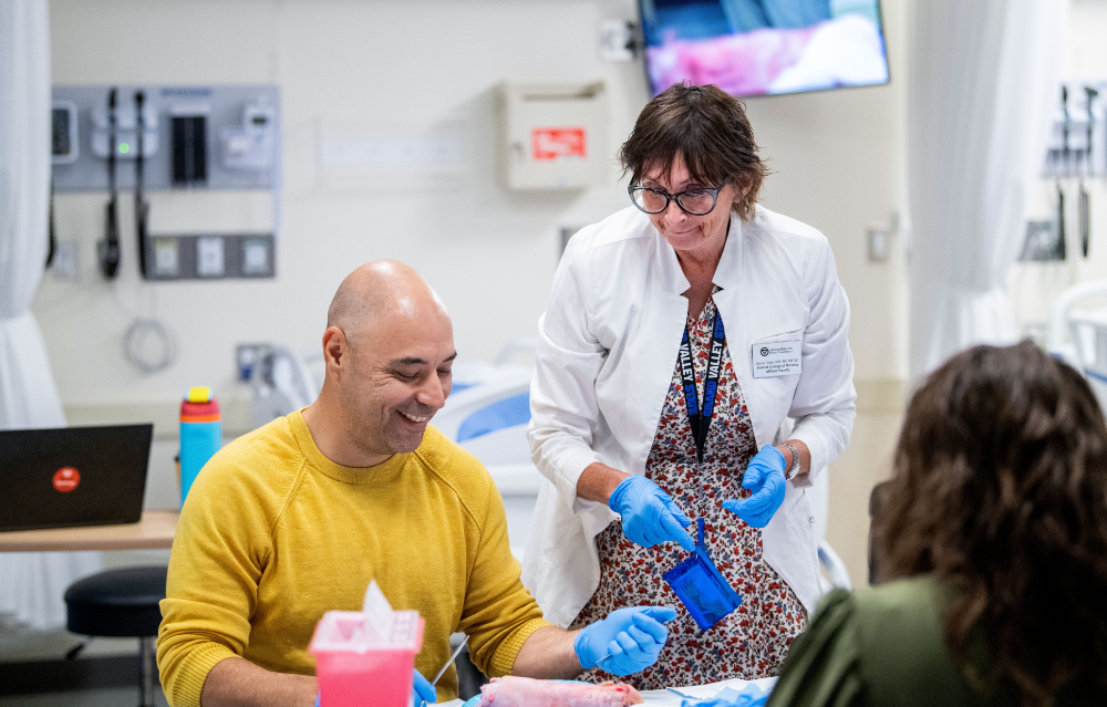 man in yellow shirt seated while nursing faculty in white coat stands over looking at table with pig's foot