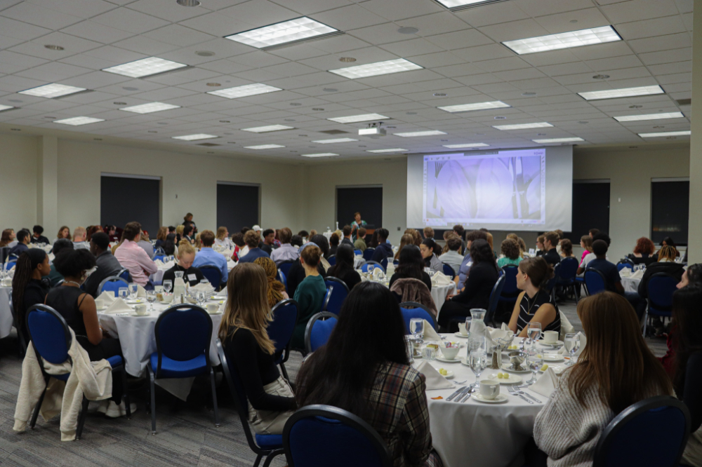 Cindy Brown up front at podium, projection screen shows place setting, students seated at round tables