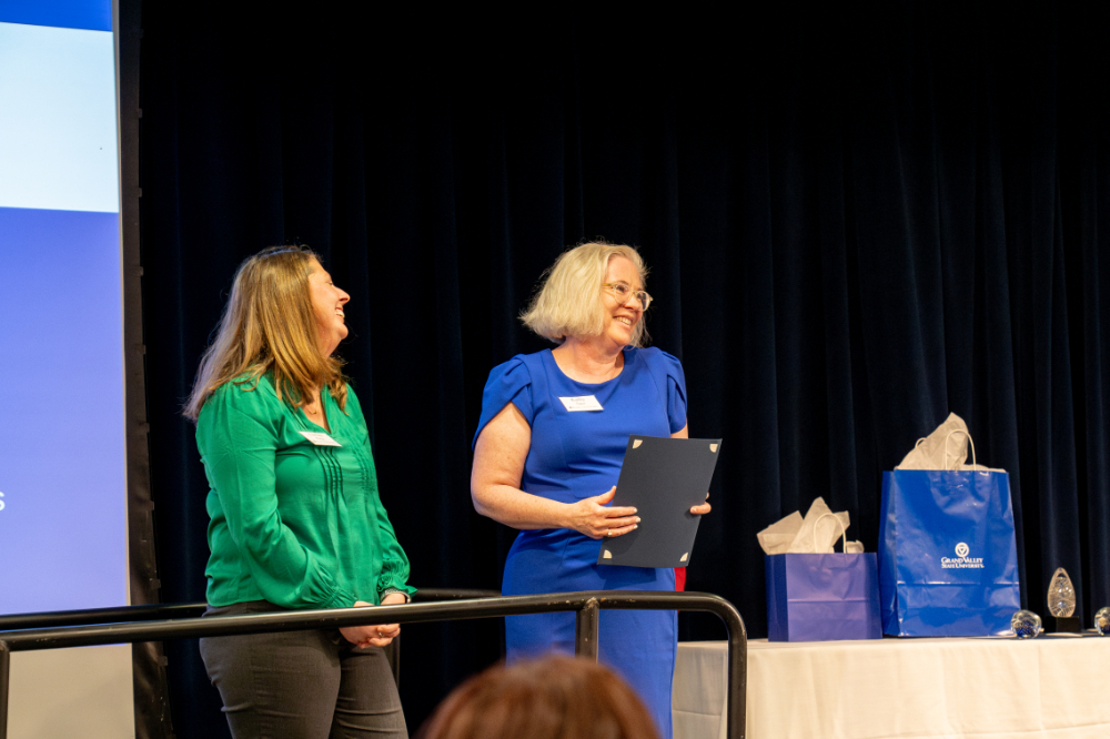 two women laugh as they stand on stage; Chelsea Ridge in green and Kathy Agee in blue dress