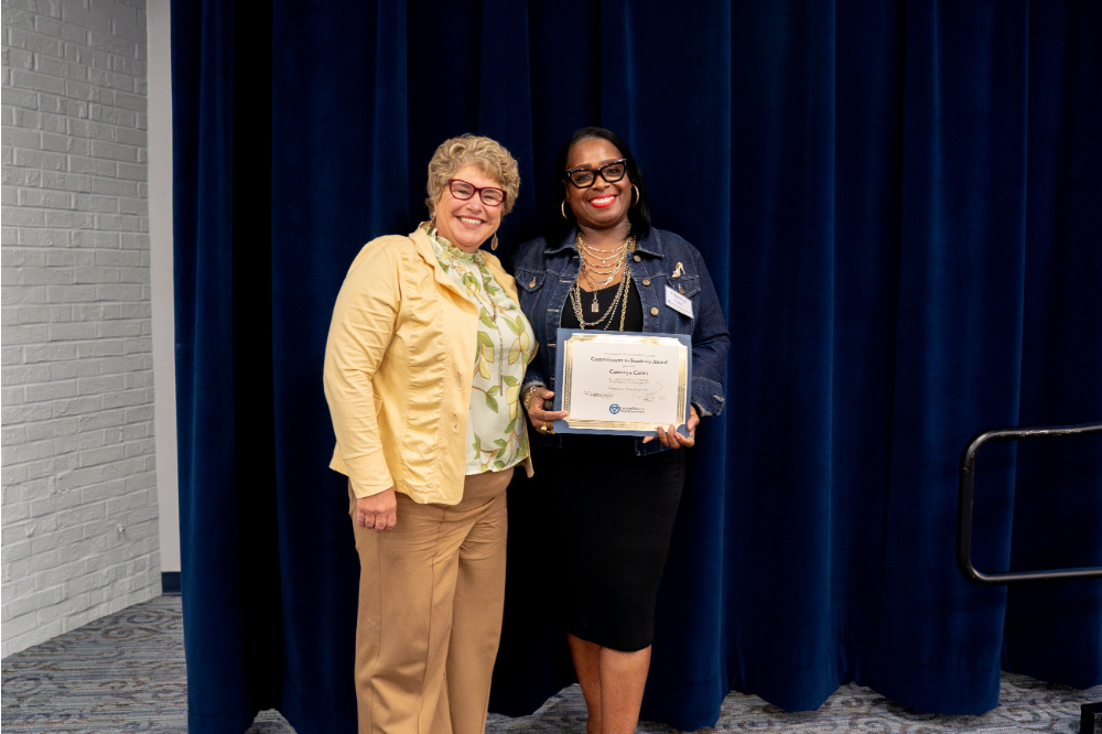 two women pose for photo in front of blue curtain backdrop; Cassonya Carter on right holds a certificate and glass award