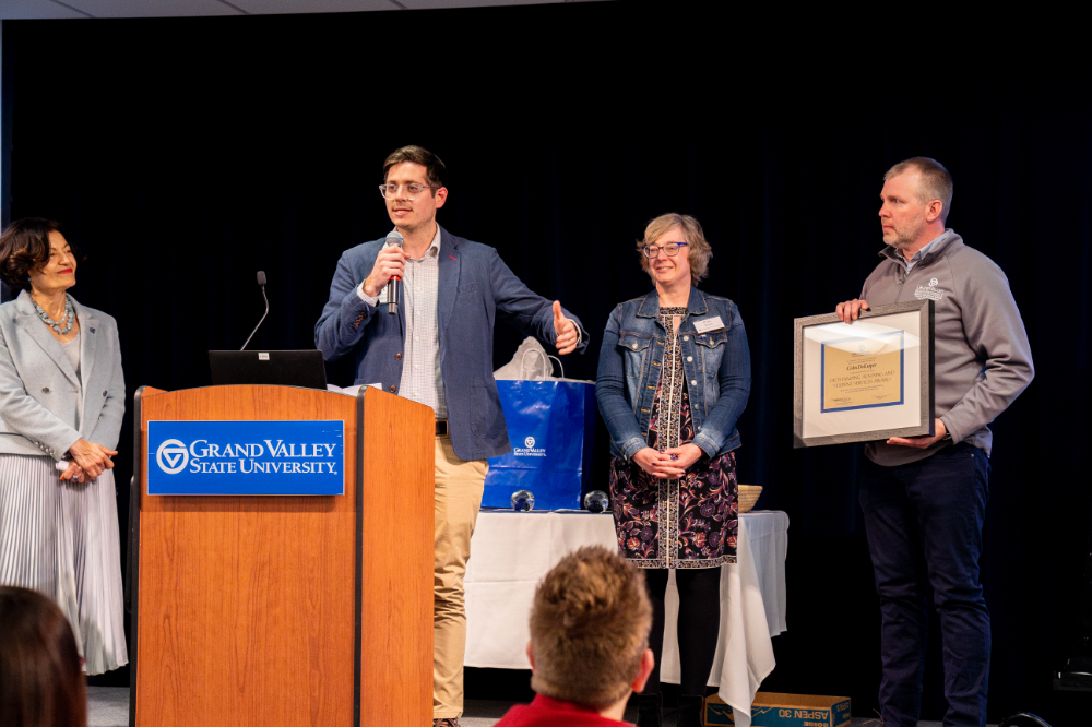 man on stage with microphone, two women on either side of him, while Colin DeKuiper holds large certificate on right of stage