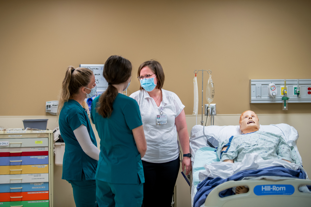 a high-fidelity manikin is in a hospital bed while two nurses in green scrubs and a woman in street clothes talk on the left side of the bed