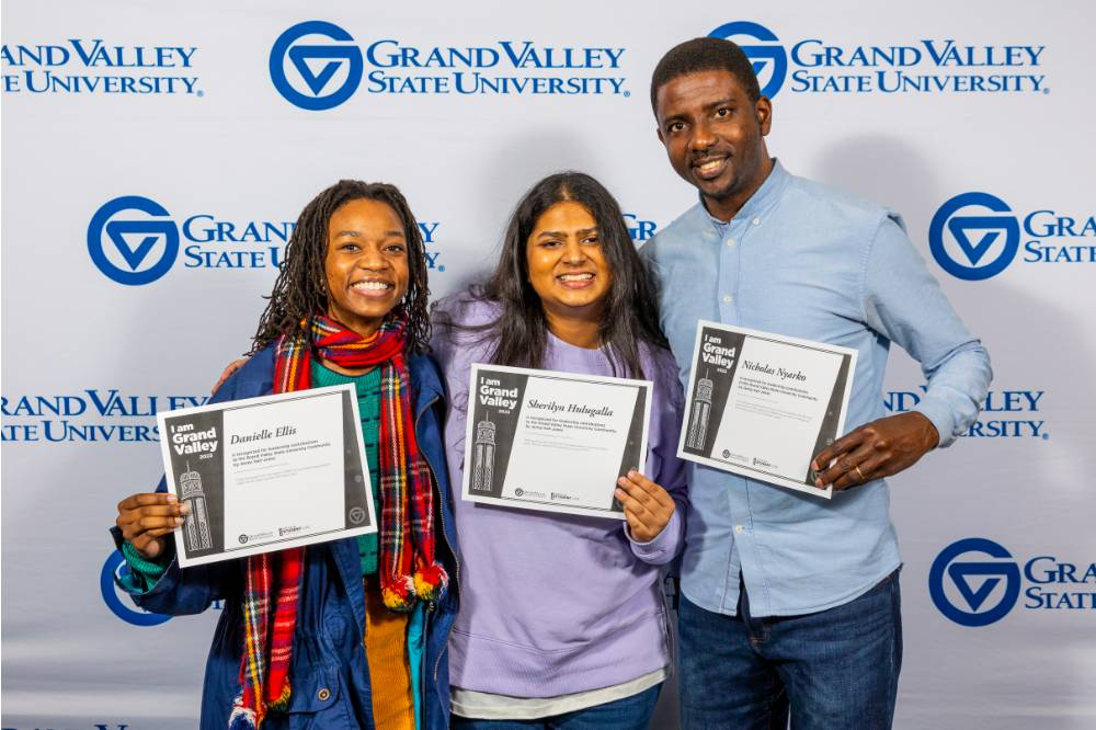 three students hold certificates in front of a background of the GVSU logo