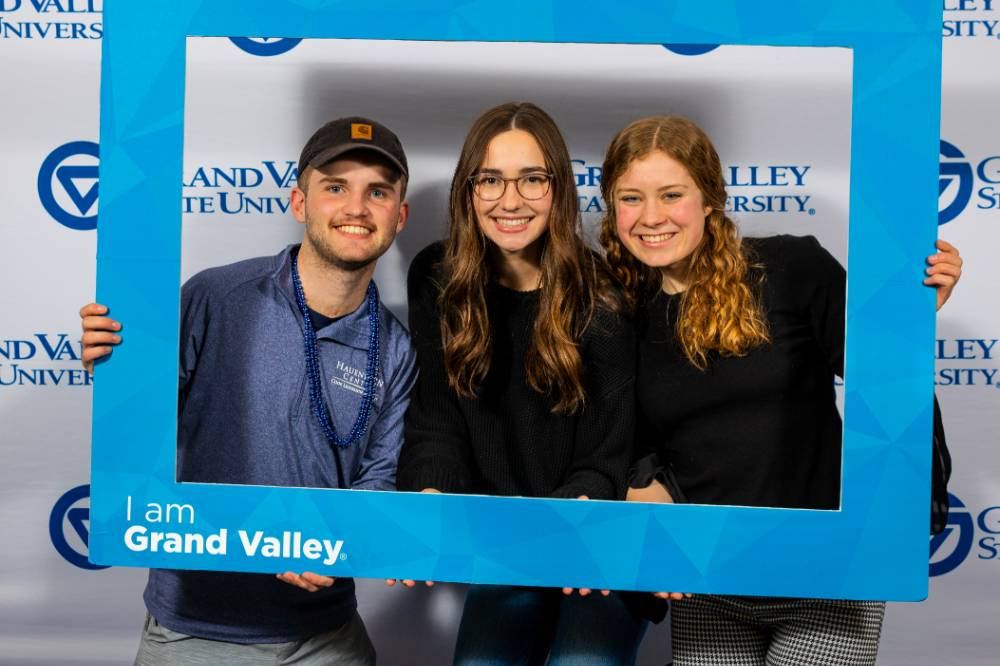 three students hold a photo prop frame with "I am Grand Valley" in the lower left corner