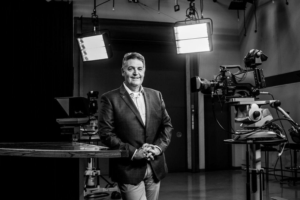 Jim Rademaker stands in the WGVU studios; two lights shine behind him, a camera is pointed at him
