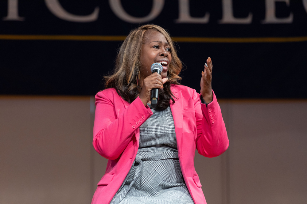 LaTosha Brown seated on stage with microphone; dressed in pink jacket and gray dress