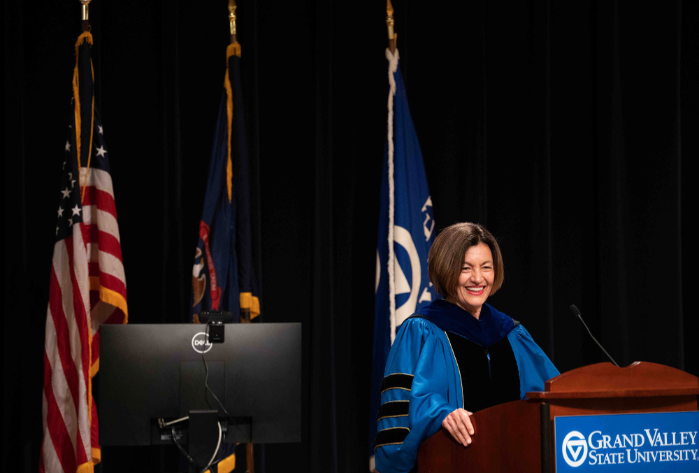 Provost Mili at the podium in front of three flags