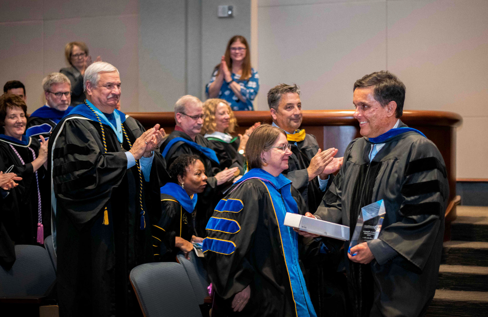 man in academic regalia holds an award while others stand and applaud
