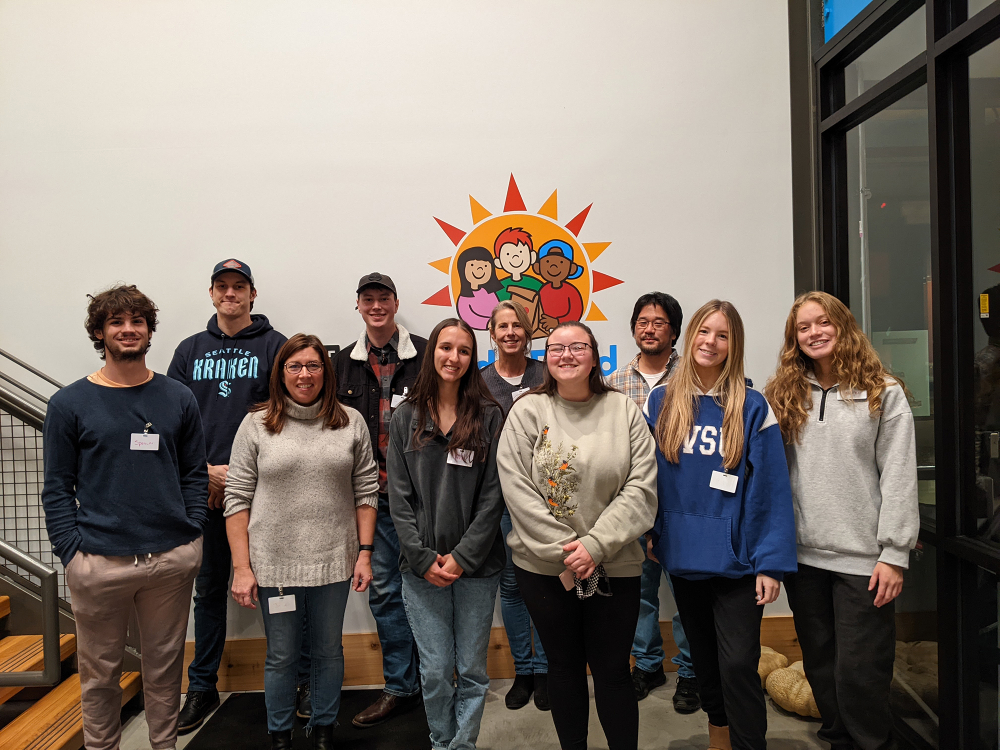 group of people stand in two rows at the base of a staircase, a logo from Kids' Food Basket is behind them on the wall