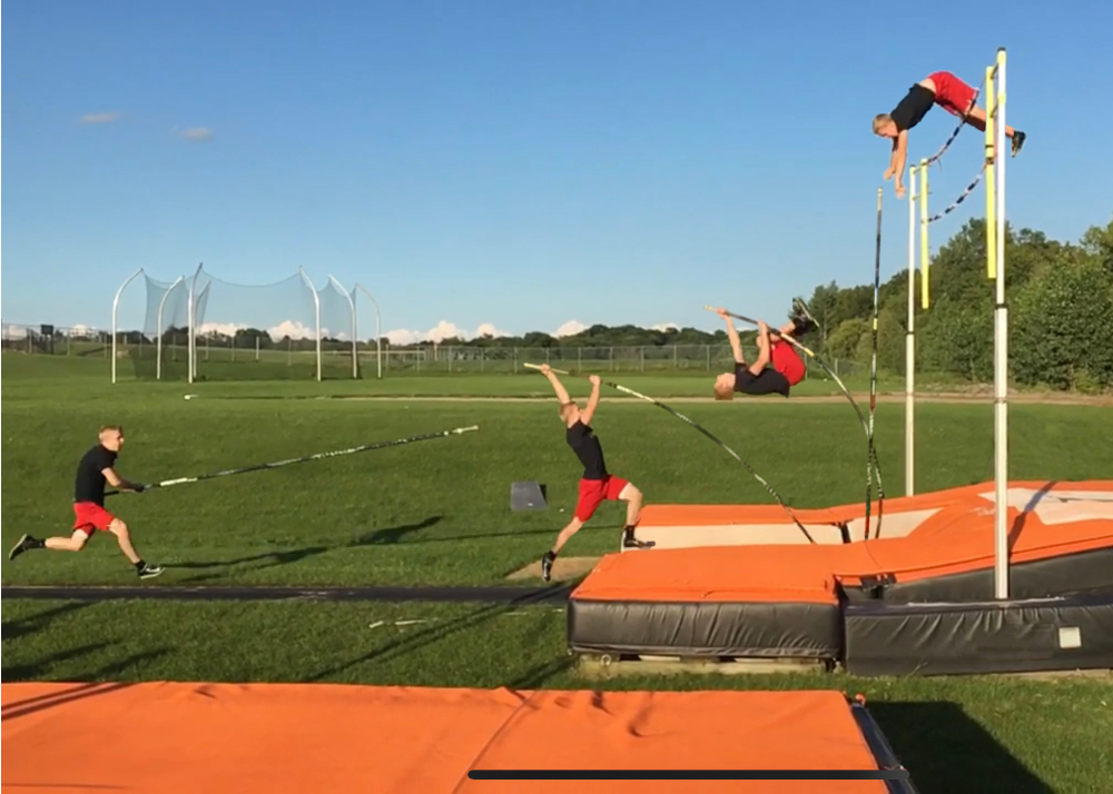 time lapse image of a pole vaulter, dressed in red shorts and black t-shirt, vaulting over a pole outside; the track mats under the pole and on the ground are orange