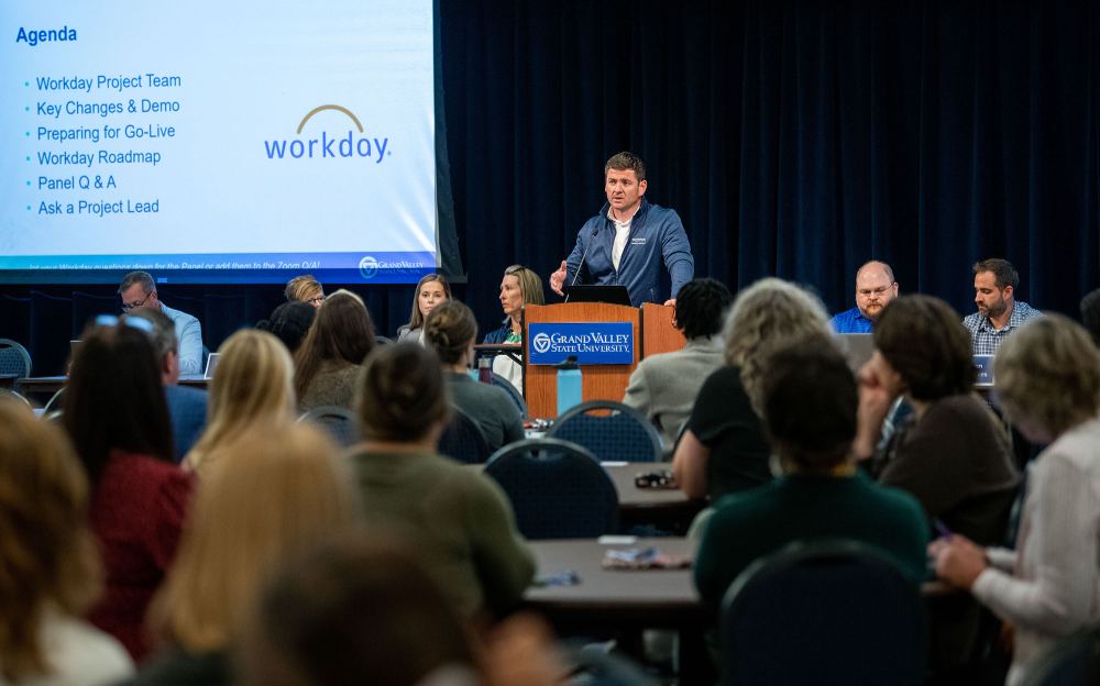 man at podium flanked by long table of people seated, audience of people at round tables look on; Workday information on projected screen behind podium