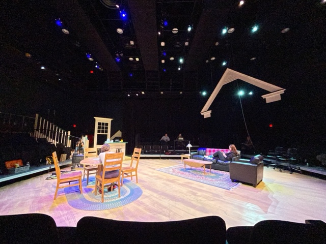 a stage during rehearsal with a piece of roof suspended on wire on the right, table and chairs on the left