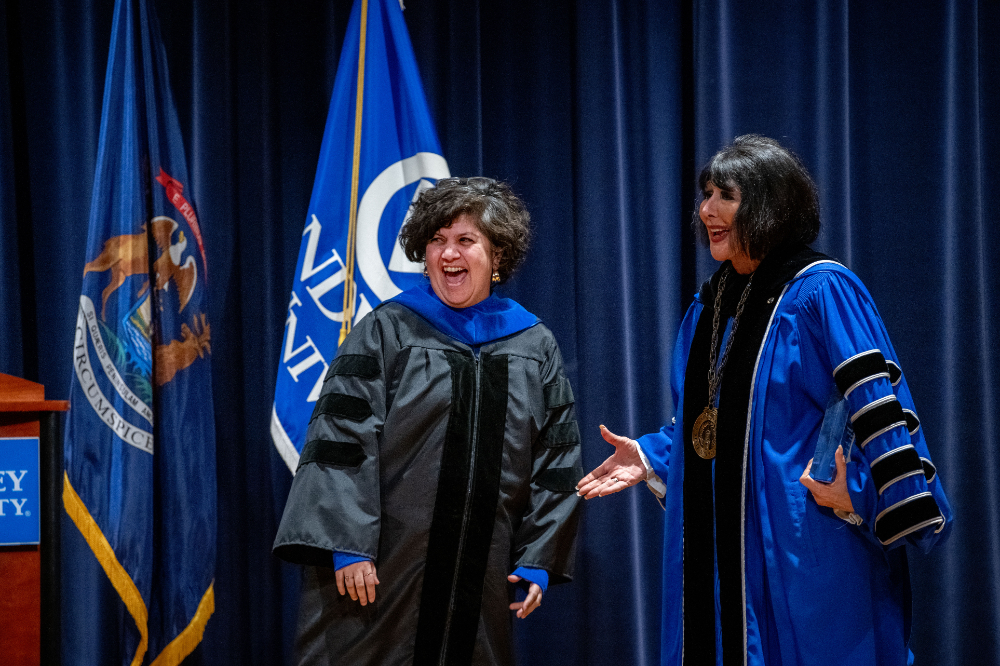 at left Samhita Rhodes and President Mantella stand on stage with blue curtain behind them and state of Michigan and GVSU flags