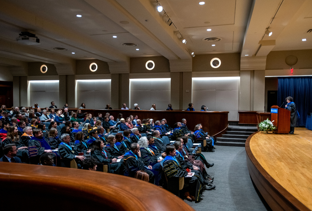 Samhita Rhodes at podium, rows of faculty in academic regalia seated in front