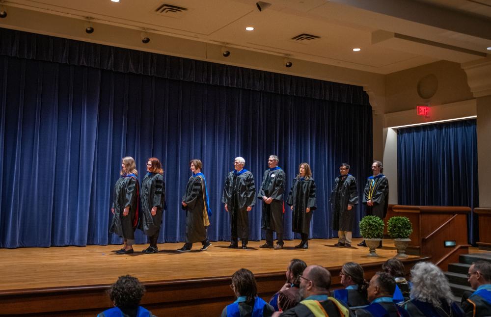 eight faculty members in academic regalia lined up on stage, blue curtain behind them