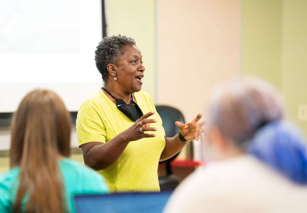 Faye Richardson-Green addresses a group of students, she is wearing a yellow dress with black mask hanging from her neck