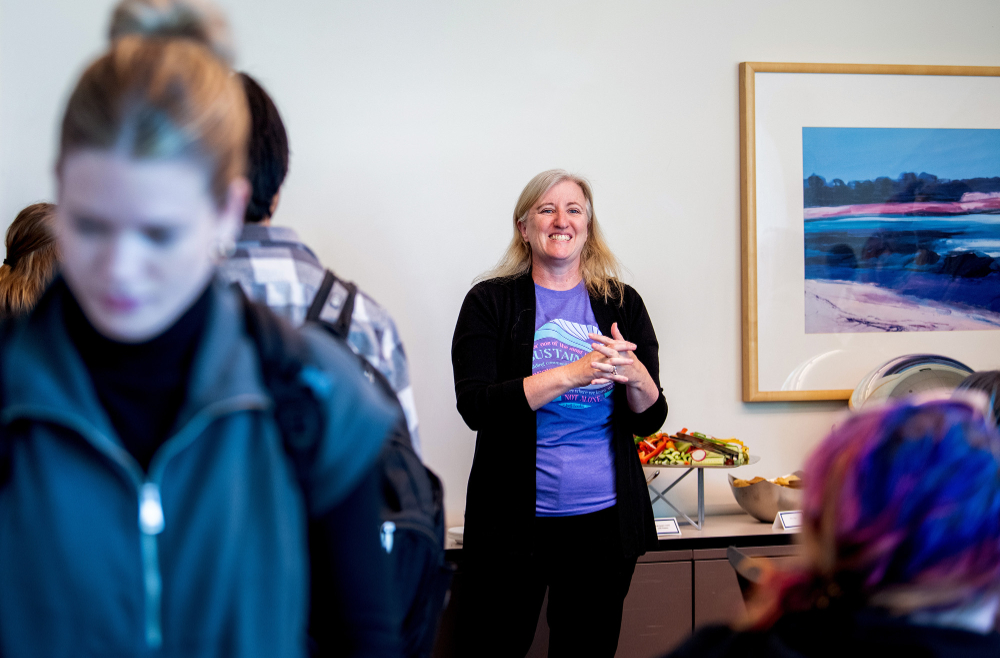 woman in black sweater stands in back of room as students mill at a buffet table