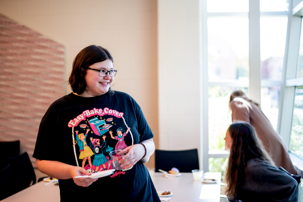 student in black t-shirt stands holding a small paper plate, students seated at table behind her