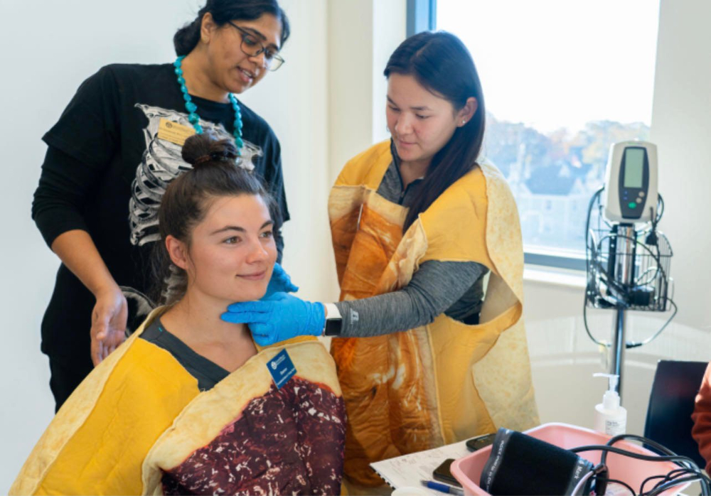 one person seated in medical lab, two people standing, one with gloved hand on seated person's vocal chords