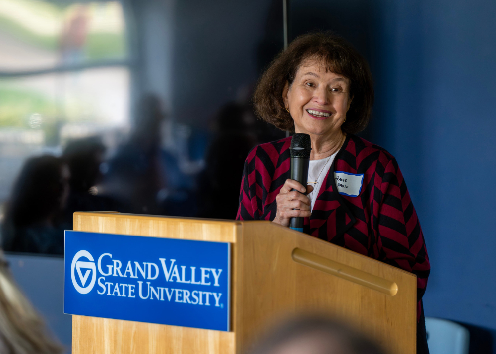 Gayle Davis holds microphone and speaks from podium with GVSU logo