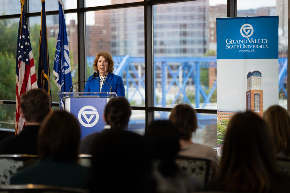 Sue Jandernoa speaks at a podium in the Eberhard Center, blue bridge in background through windows