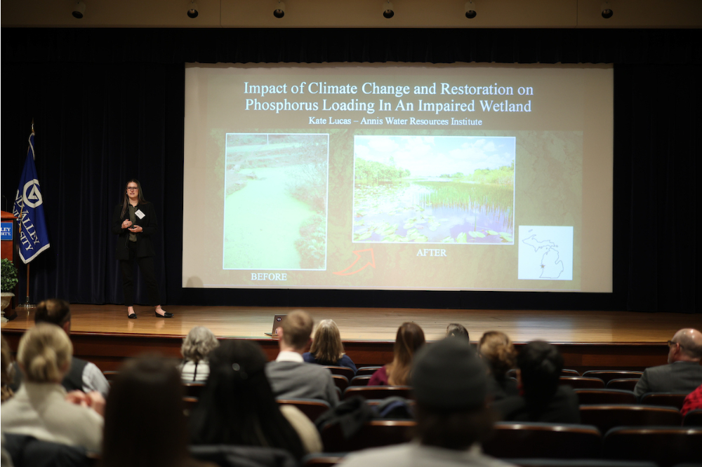 Kate Lucas  stands on stage next to a projection screen with her research slide on it.