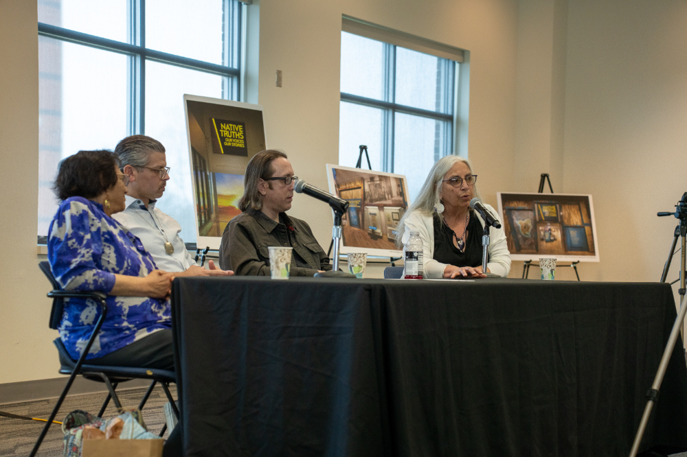 four people seated at table with microphones in front of them, posters on display behind them