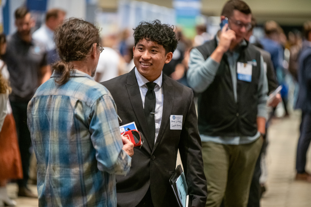 student in suit and tie smiles at a person with his back to the camera; a man in the background is holding a cell phone to his right ear and speaking