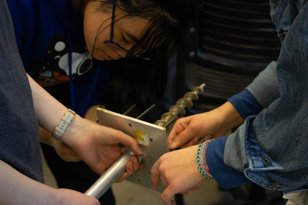three people holding a mechanical device; two sets of hands on this metal while one student is seen on the left