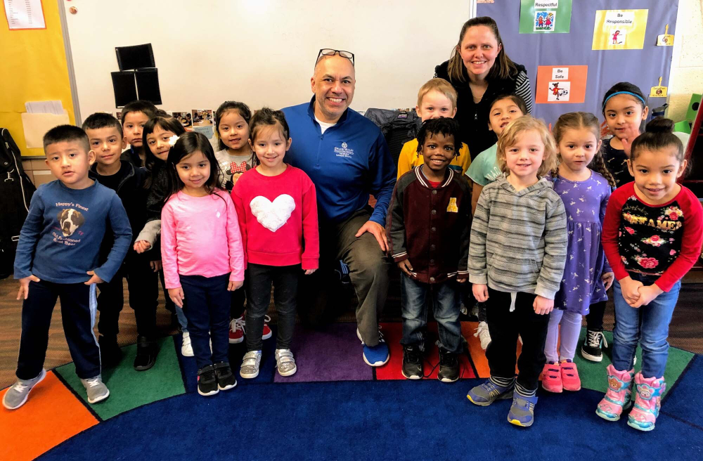 man kneels on floor behind two rows of elementary school children, teacher also in back row