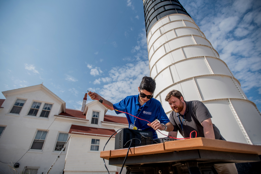 in front of a lighthouse, two people work at a table fixing wires into a ham radio