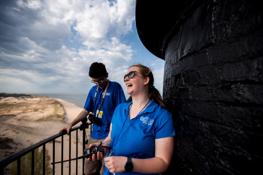 two people holding a wire on a balcony of a lighthouse with sand dunes and Lake Michigan in background