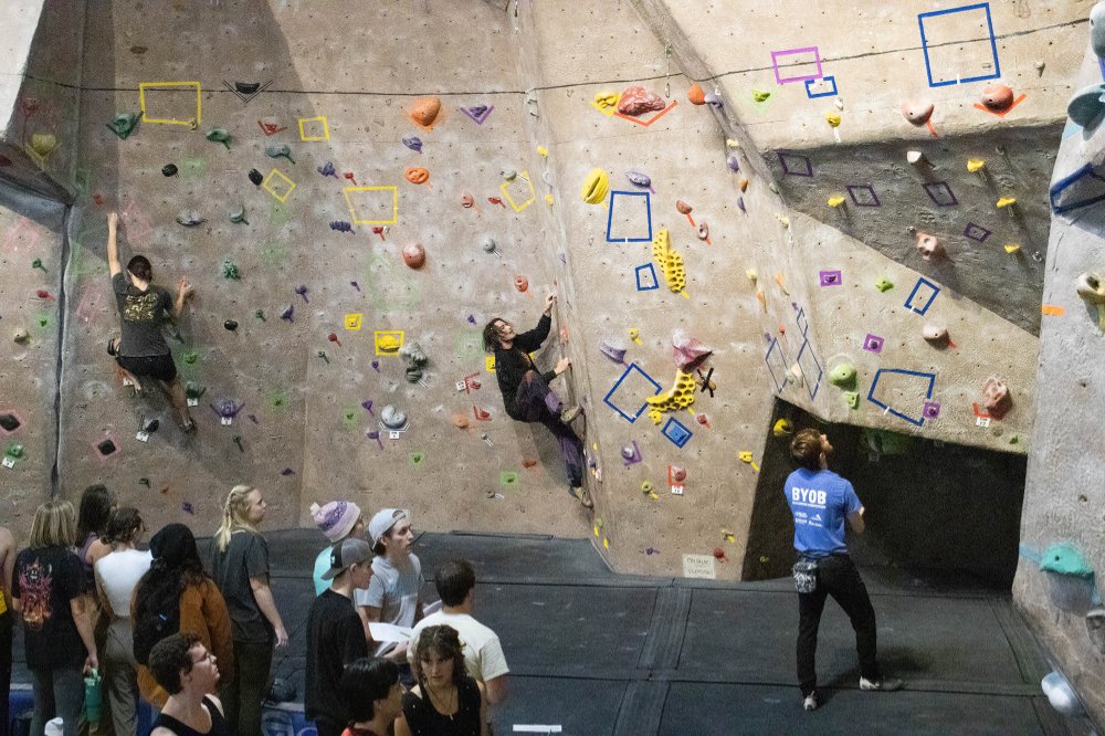 Two people on climbing wall, one person watching from below in blue shirt; crowd at left