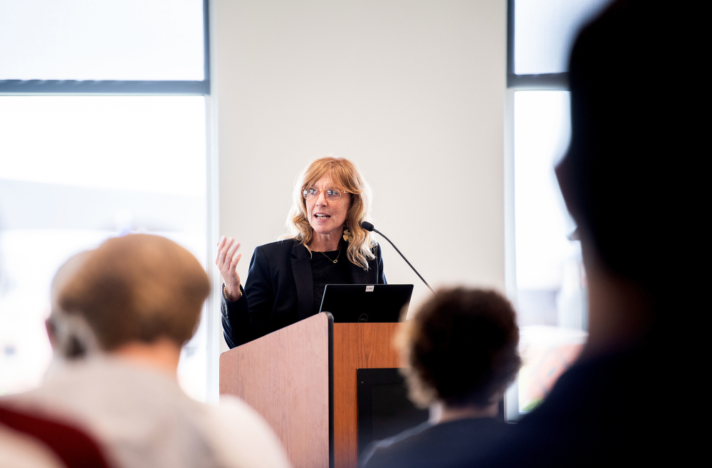 woman standing behind podium, right arm raised while talking to seated audience
