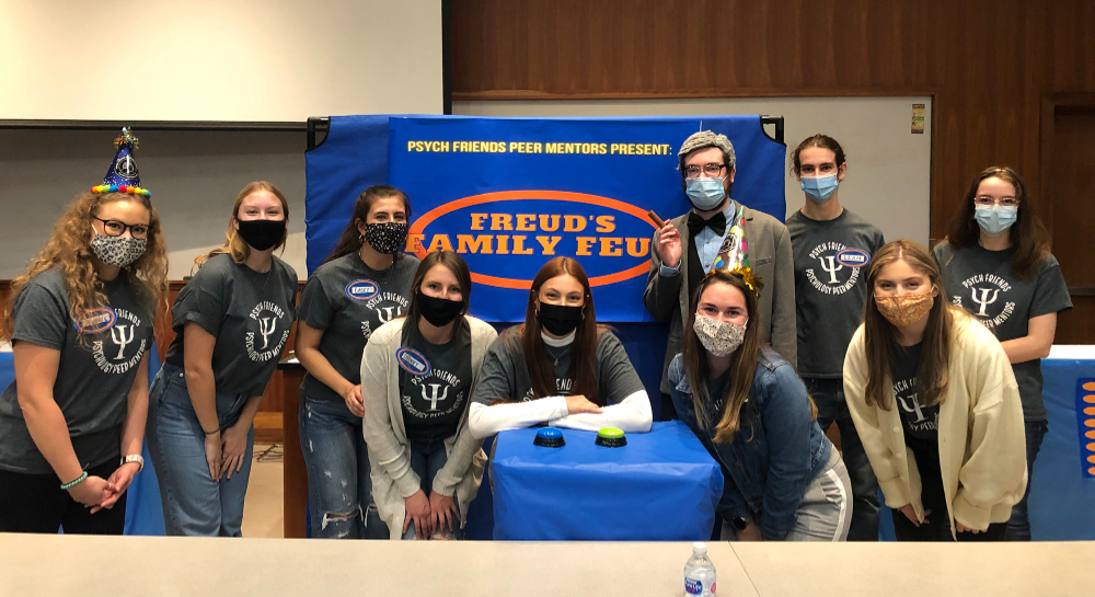 10 people standing behind podium and in front of banner, Freud's Family Feud. They are all wearing masks.