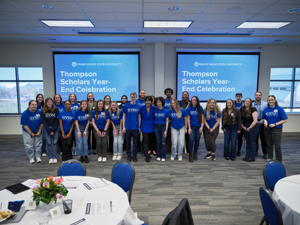 large group of students in blue tshirts stand with president Mantella in front center in front of screens that read Thompson Scholars Year-end celebration