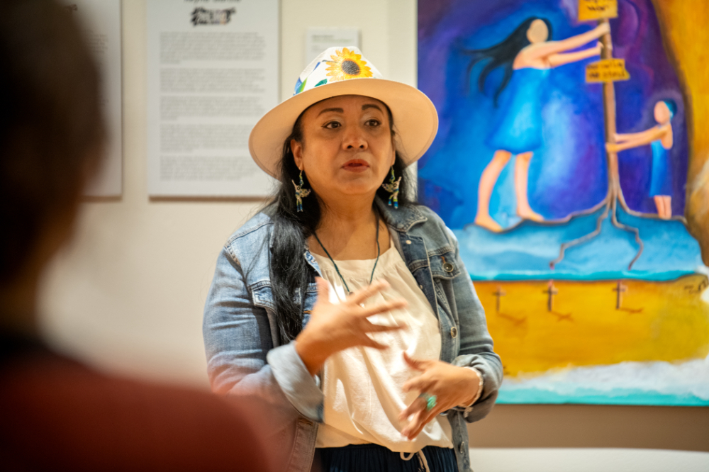 Reyna Garcia in hat with sunflower stands in front of colorful painting