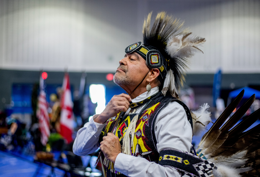 a participant at the pow wow adjusts his clothing, including a headdress and beaded vest in the Fieldhouse