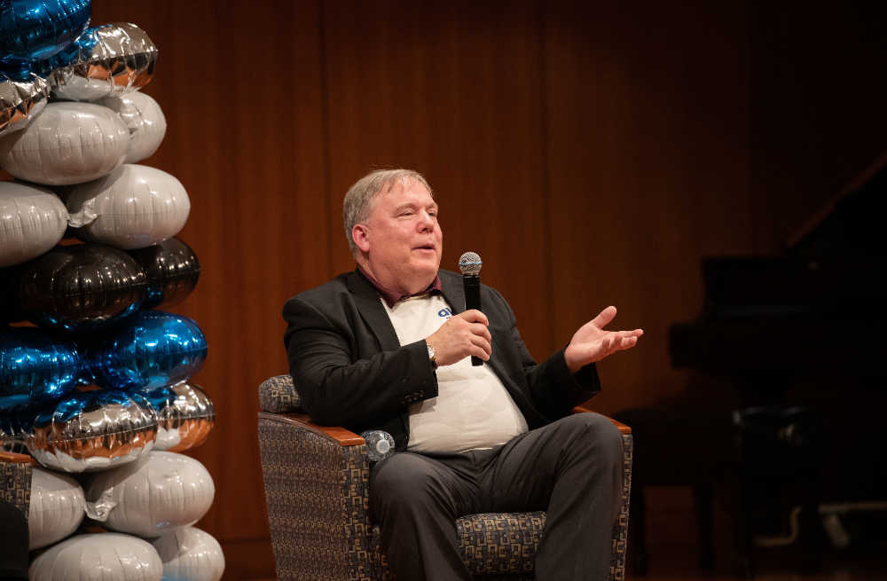 Andy Beachnau, seated in chair with microphone, gestures with hand. A balloon arch is at his right