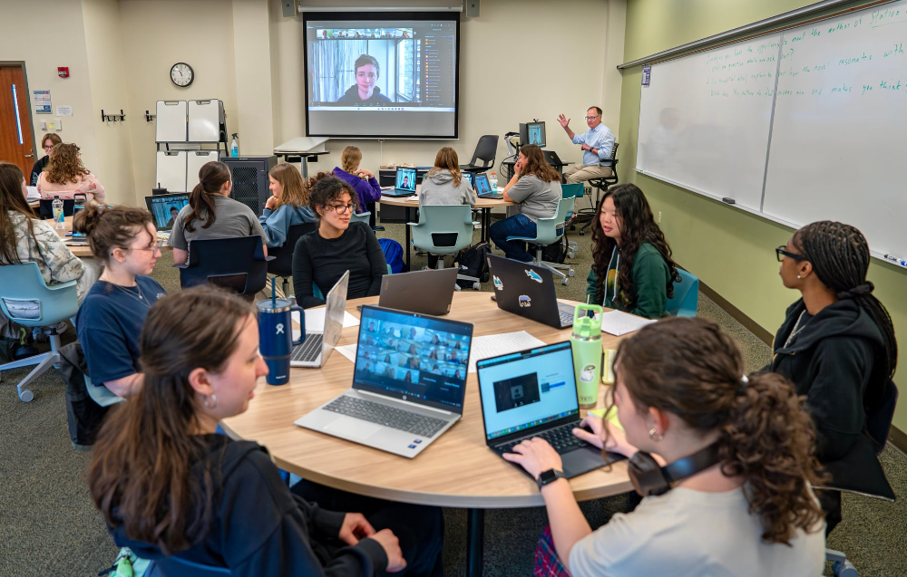 Emily St. John Mandel on large projection screen speaking to a class of students at round tables with laptops out, David Eick is at back right with arm raised