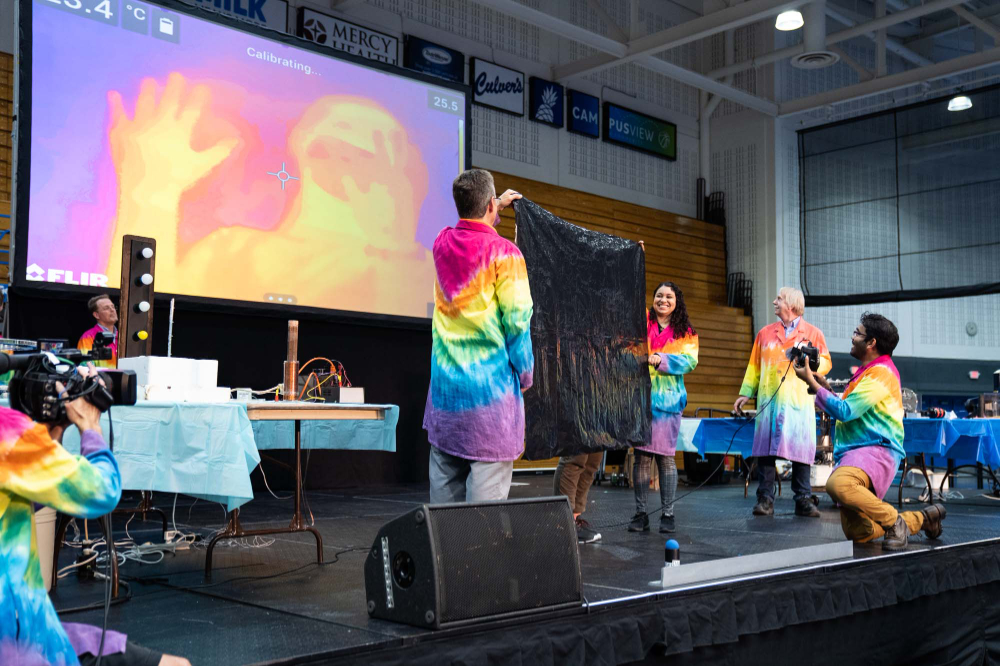 thermal image on screen in back, two people in rainbow lab coats holding black sheet, other person in front with camera, videocamera person on left