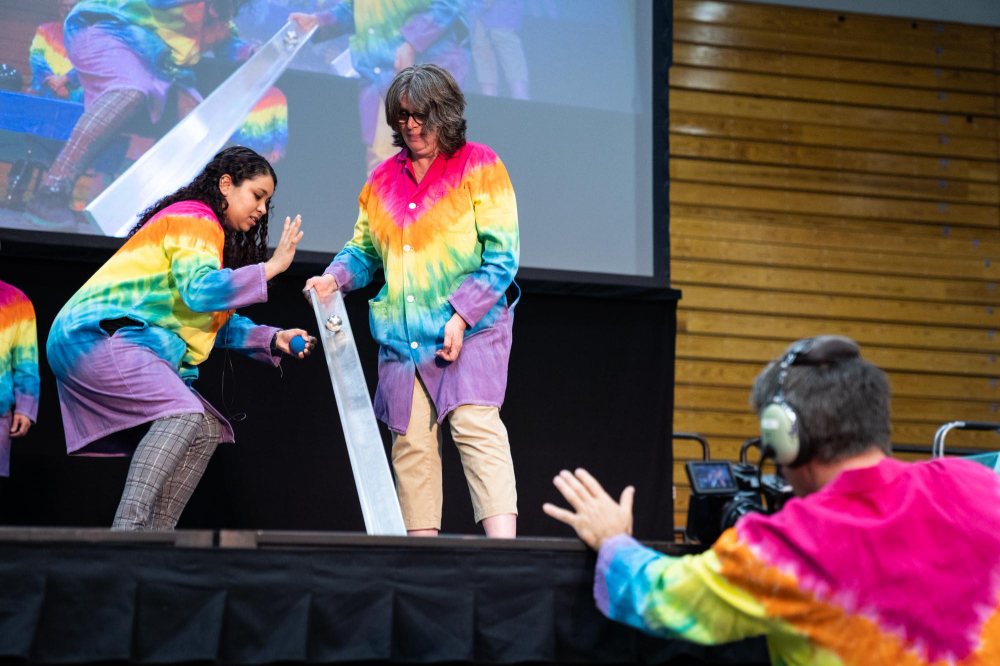 two people in rainbow lab coats roll metal balls down a metal tube; a videographer is in front, they are projected on the screen in back