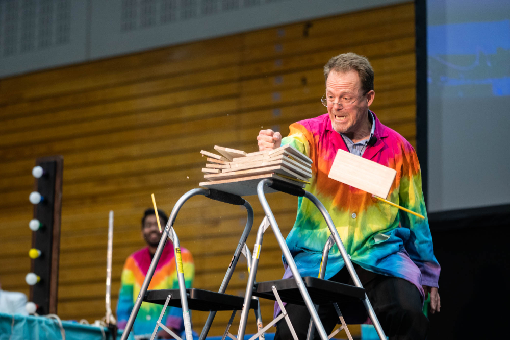 man in colorful lab coat breaks what looks like plywood with his fist