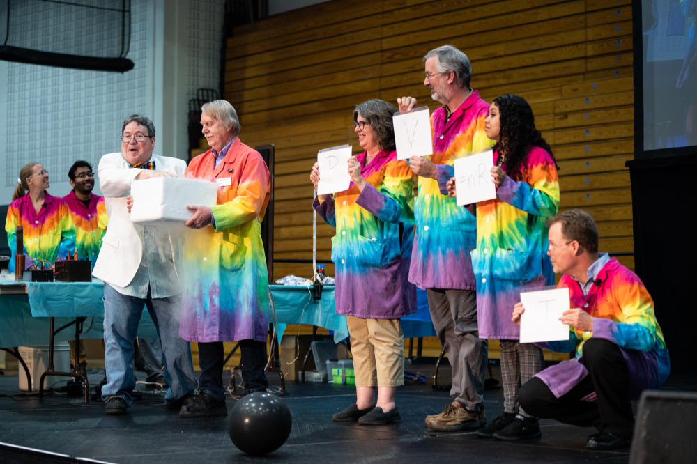 large group on stage holding signs, man at end talks about a box held by another man in a rainbow labcoat