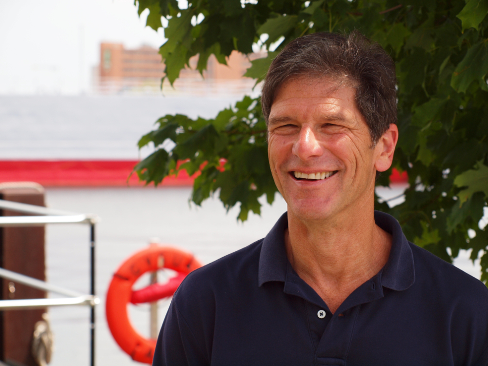 Alan Steinman standing in blue polo shirt outside near shoreline and in front of a tree