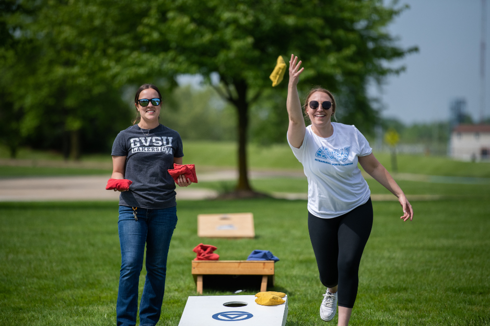 two people standing and playing cornhole with GV themed base; woman on right throwing yellow bag