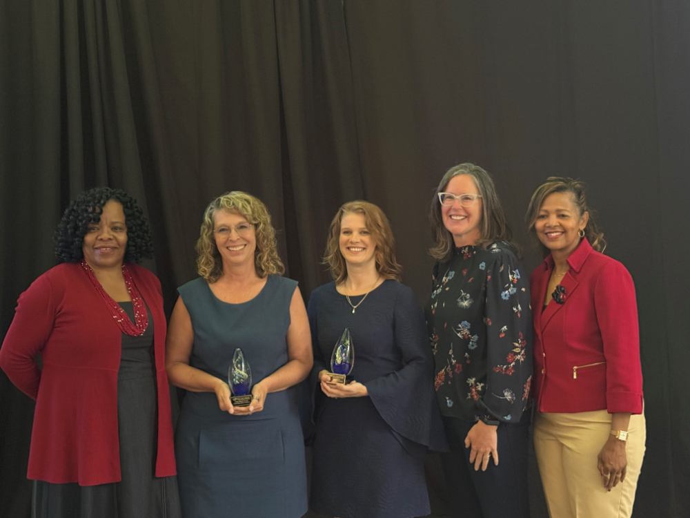 five people standing in a row, two women holding glass trophies are second and third from left
